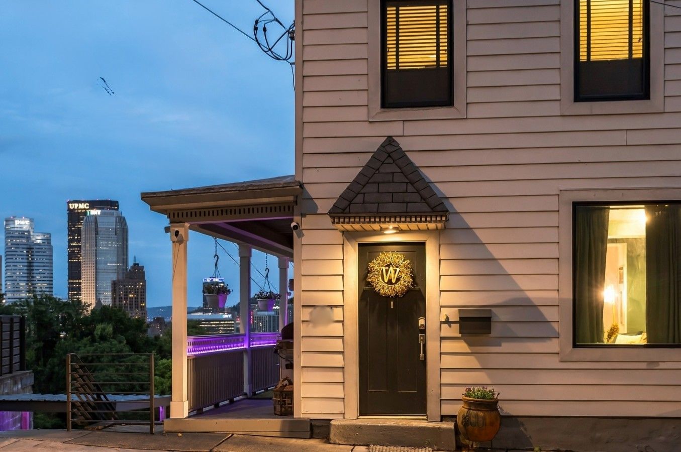 A two-story building with a black door featuring a gold emblem, a porch with purple lighting, and a city skyline in the background.