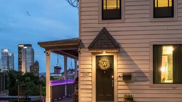 A two-story building with a black door featuring a gold emblem, a porch with purple lighting, and a city skyline in the background.