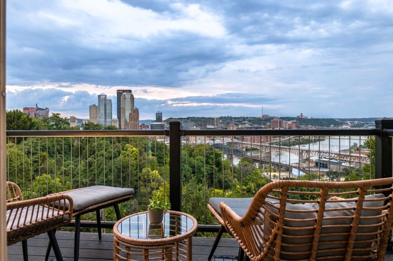 A balcony with wicker furniture, a glass table, and a view of a cityscape and river.