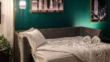 A neatly made bed with a white duvet and two rolled towels, a grey sofa, a floor lamp, and two cityscape paintings on teal walls.