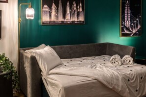 A neatly made bed with a white duvet and two rolled towels, a grey sofa, a floor lamp, and two cityscape paintings on teal walls.
