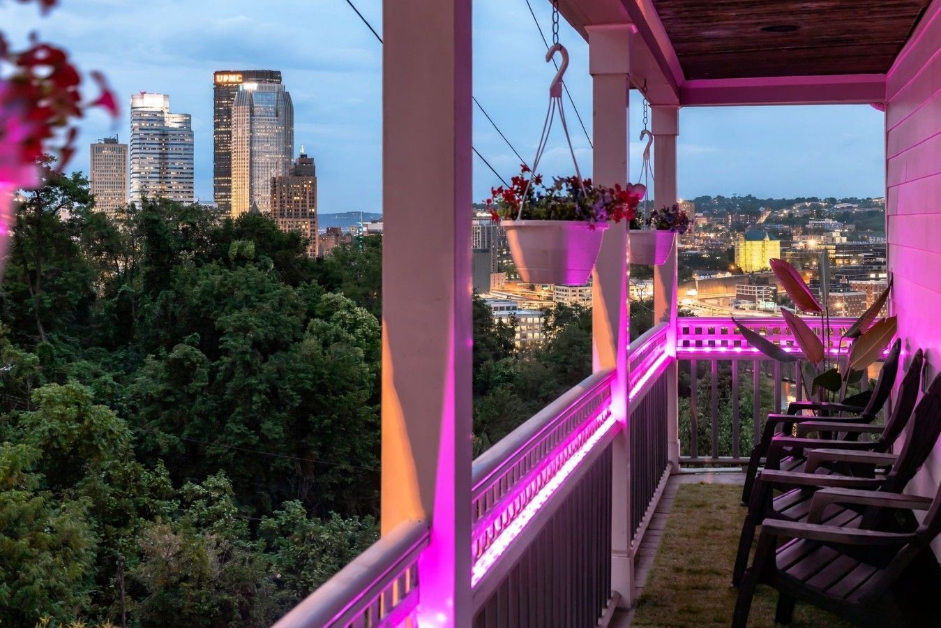 A balcony with hanging flower baskets, a railing, and a view of a cityscape at dusk.