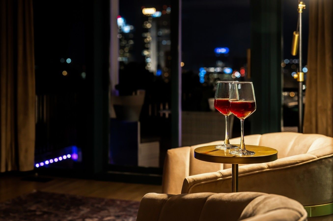 A room with a view of a cityscape at night, a leather armchair, and two glasses of red wine on a small round table.