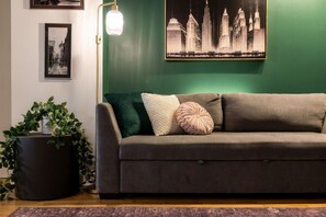 Una habitación con pared verde, un sofá gris con cojines, una lámpara de pie y un cuadro enmarcado de un paisaje urbano.