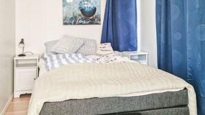 Siististi pedattu sänky, jossa on raidallinen vuodevaatteisto, harmaa verhoiltu pääty ja tyyny, jossa on teksti ”Balance”. Sängyn vieressä on yöpöytä, jossa on lamppu ja pieni valkoinen yöpöytä. Seinällä sängyn yläpuolella on taulu.