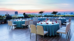 Un área de comedor en la azotea con mesas redondas preparadas para una comida, rodeada de sillas y con vistas a un paisaje urbano al atardecer.