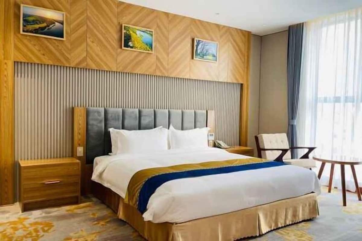 Ein Hotelzimmer mit einem großen Bett, einem Holzkopfstück, einem Nachttisch, einem Sessel und Blick ins Freie durch das Fenster.