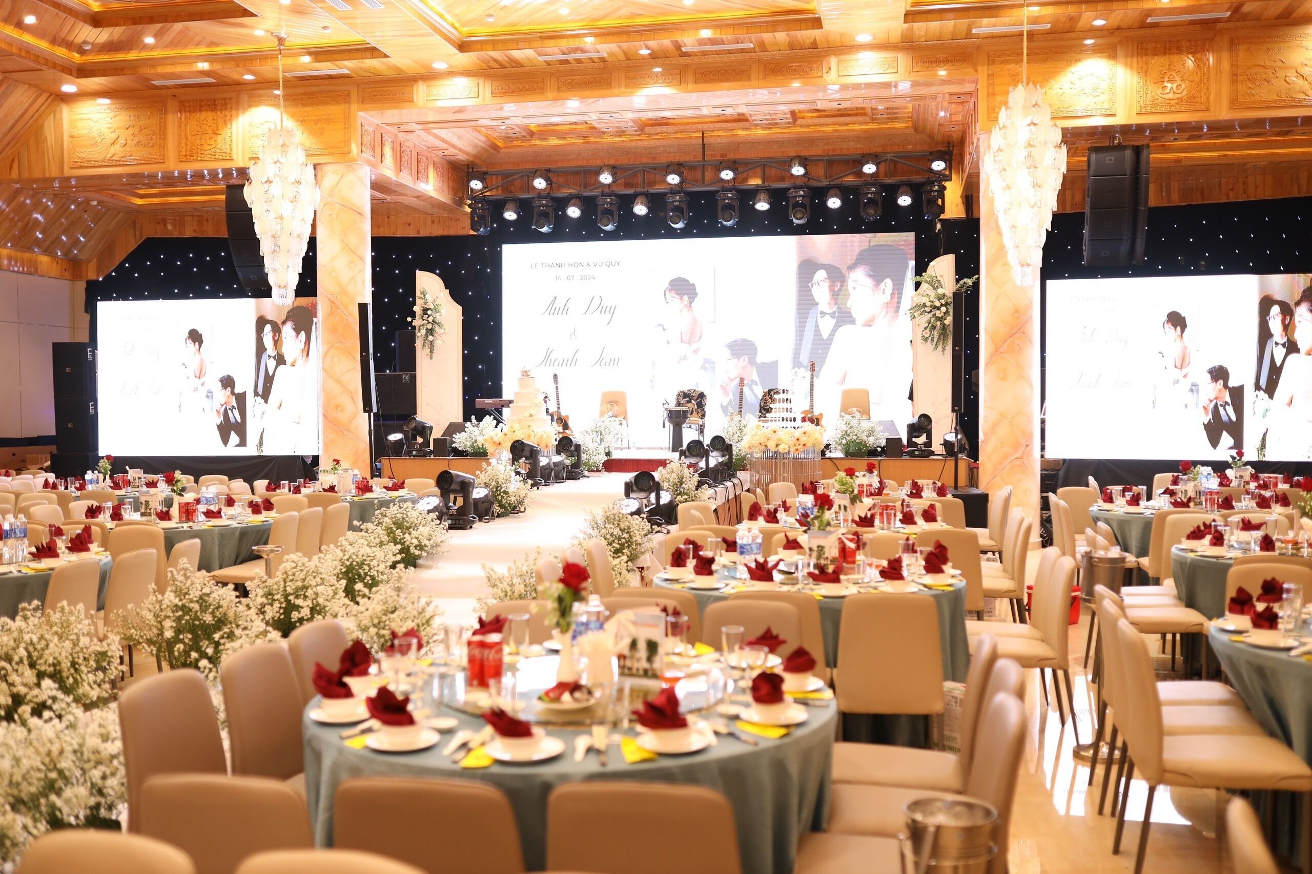 Un salón de banquetes con mesas redondas dispuestas para un evento formal, con centros de mesa florales y un escenario con una pantalla grande que muestra imágenes.