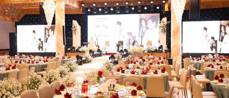 Un salón de banquetes con mesas redondas dispuestas para un evento formal, con centros de mesa florales y un escenario con una pantalla grande que muestra imágenes.