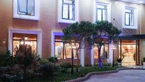 Entrada de hotel con jardín ajardinado, ventanas amplias y fachada bien iluminada.