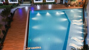 Una piscina con un deck de madera que la rodea, un área de descanso con una silla y un pequeño jardín con plantas.
