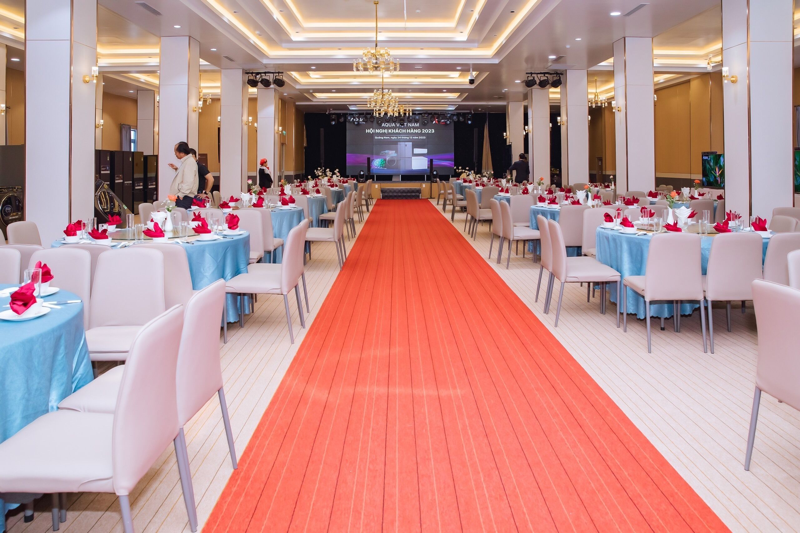 A banquet hall with a central stage, rows of tables with blue tablecloths, and a red carpet.