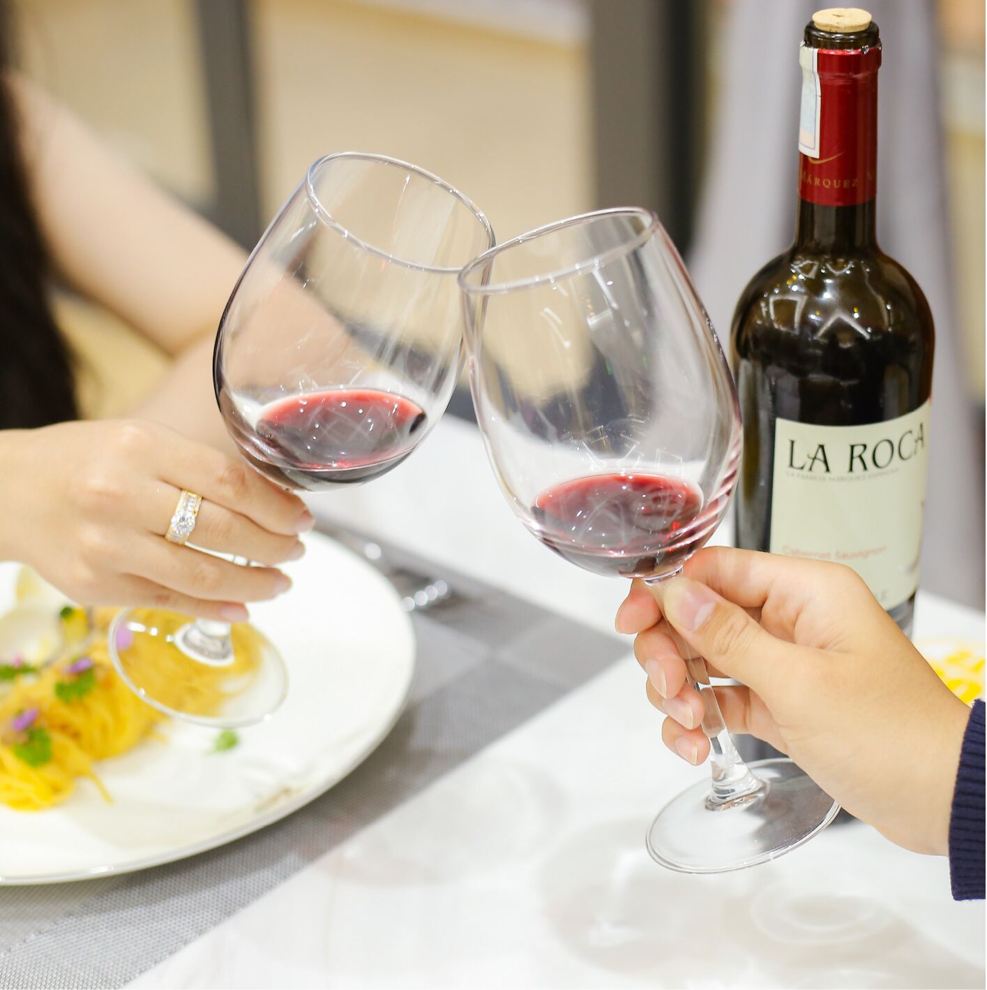Dos personas sostienen copas de vino con una botella de vino La Rocca al fondo.