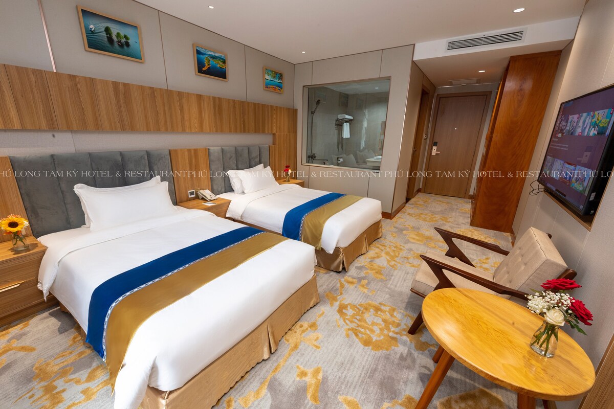 A hotel room with two beds, a wooden headboard, a large mirror, a wooden table with a vase, and a carpeted floor.