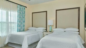 A hotel room with two beds, a nightstand with a lamp, and a window with sheer curtains.