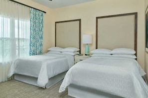 A hotel room with two beds, a nightstand with a lamp, and a window with sheer curtains.