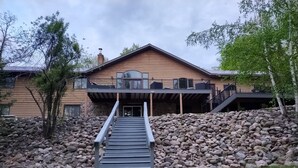 Un edificio de dos pisos con un muro de contención de piedra, una terraza de madera y una escalera que lleva a la entrada.