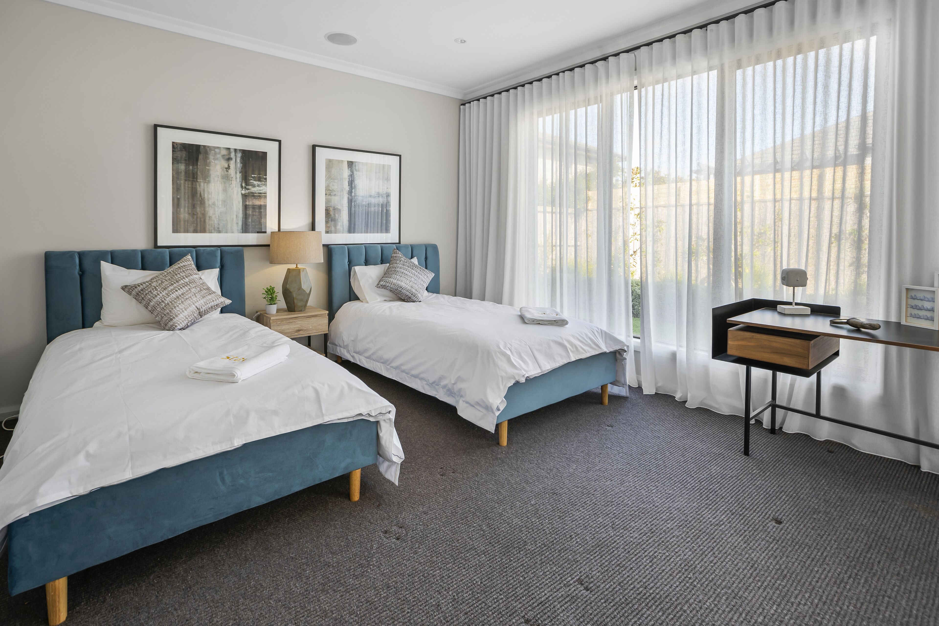 A hotel room with two beds, a desk, a chair, a lamp, and a window with curtains.