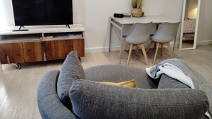 Moderni olohuone, jossa on litteänäyttöinen televisio, harmaa sohva, puinen kaappi ja pieni työpöytä tuoleineen.