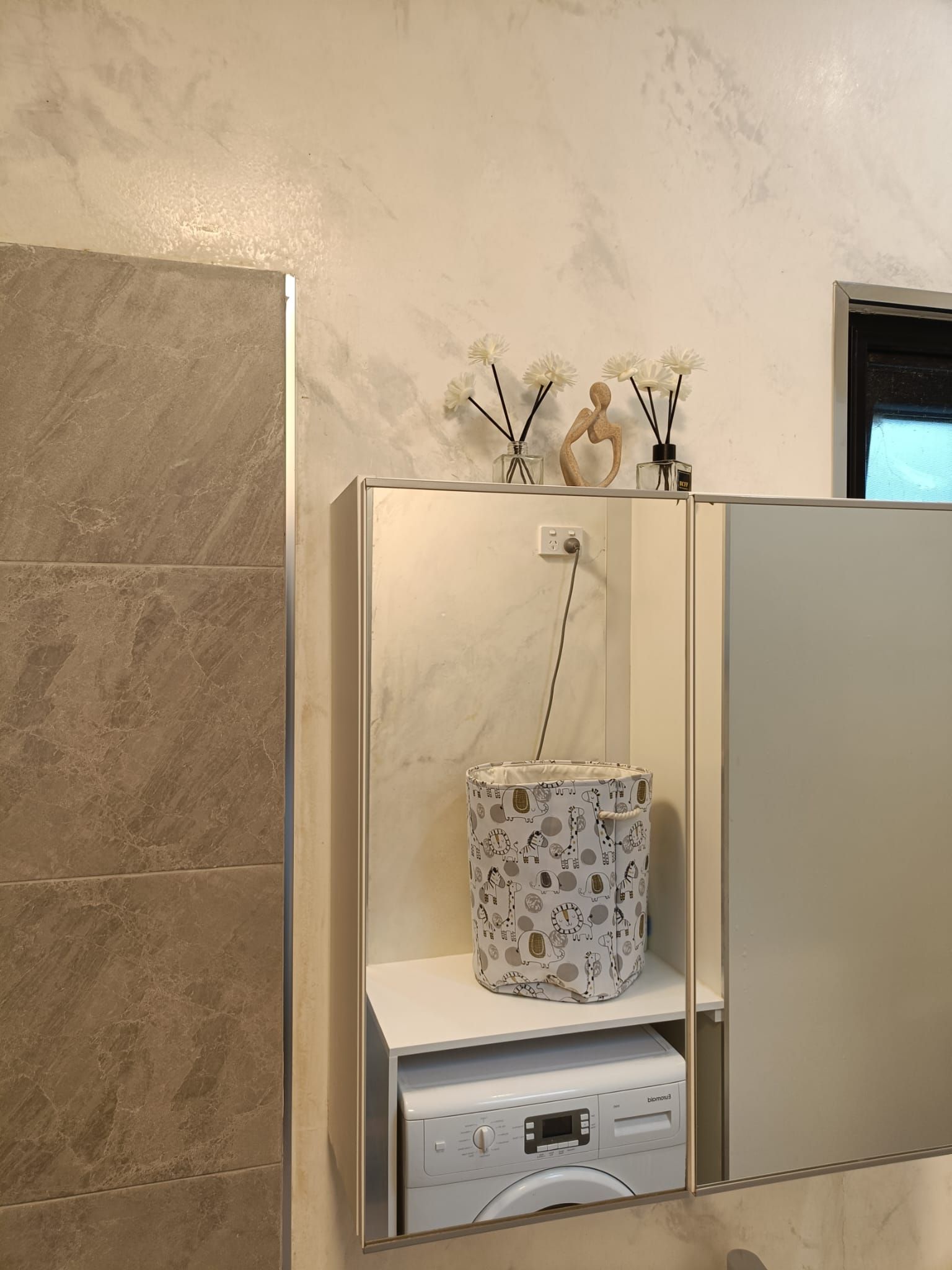 Un baño con lavadora, un armario espejo y un florero con flores en la pared.