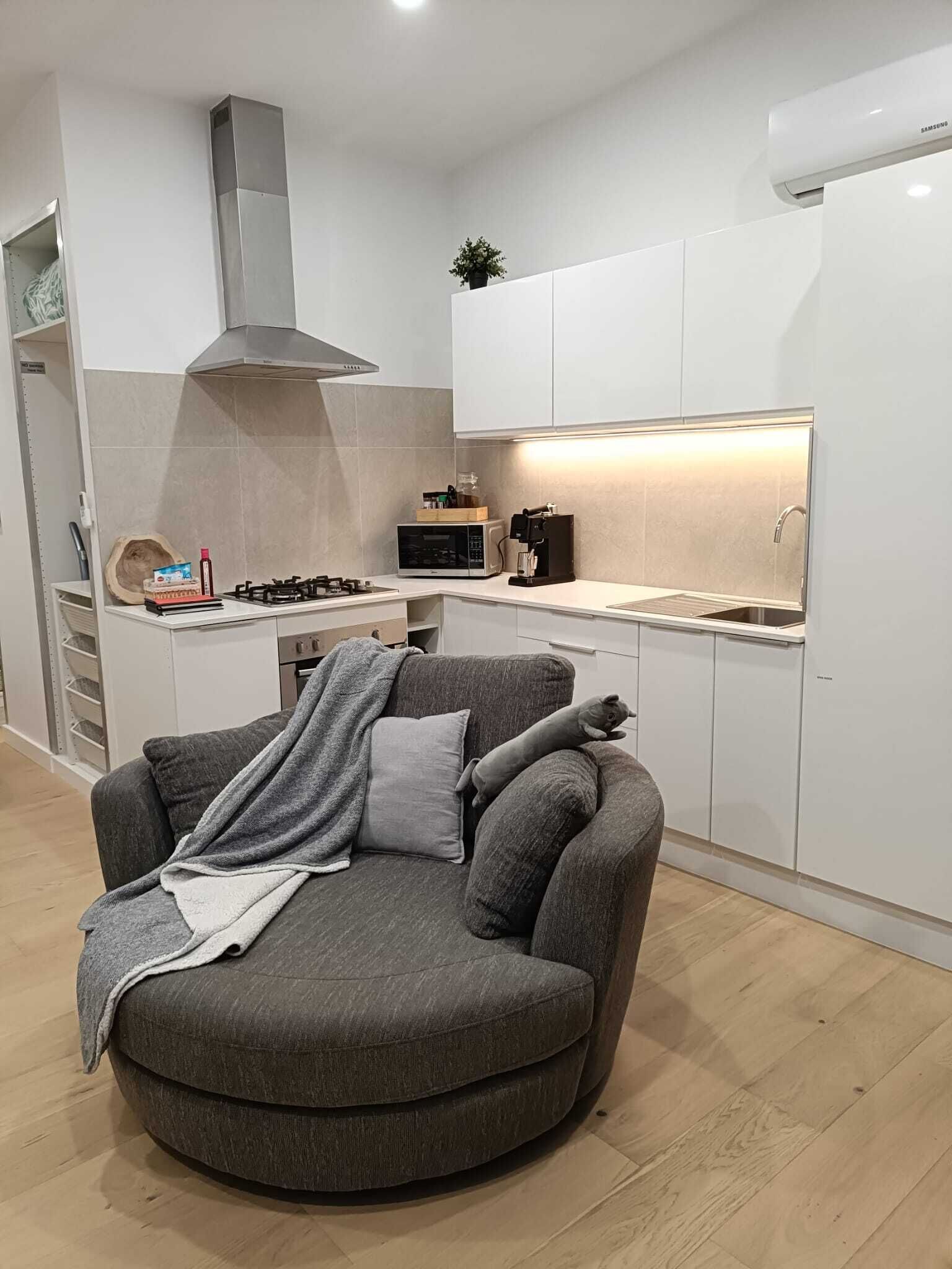 A modern kitchen with white cabinets, a microwave, and a stove. There is a grey armchair with a blanket and pillows in the foreground.