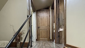 Un pasillo con puerta de madera, escalera con barandilla y un jarrón con plantas secas a la izquierda.