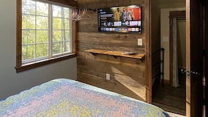 Un dormitorio con un ventanal grande, una pared de madera, una cama con colcha de colores y un televisor montado mostrando un menú.