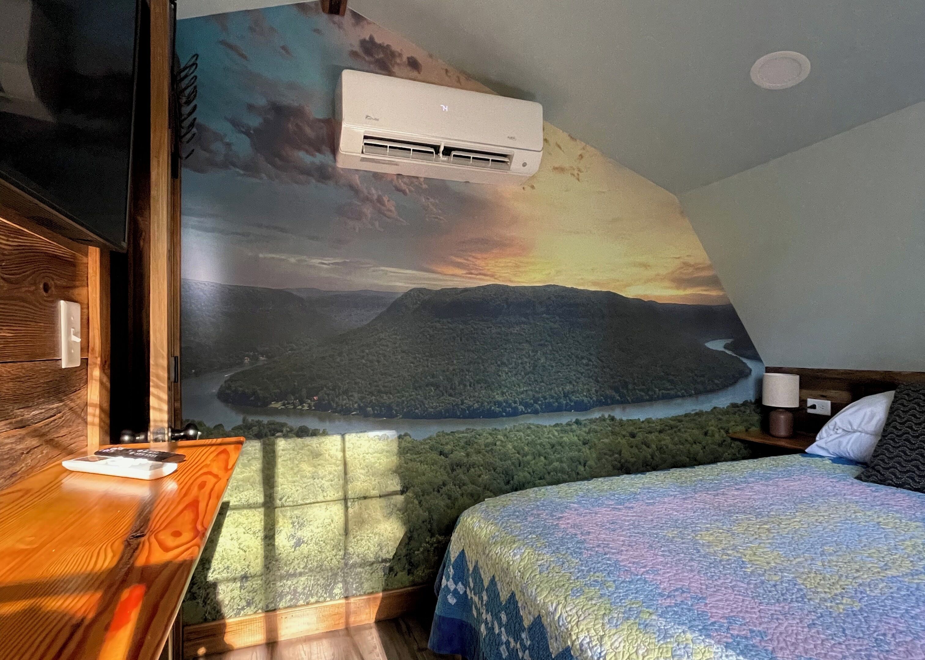 A bedroom with a mural of a landscape, a bed, a nightstand, a lamp, and an air conditioning unit.