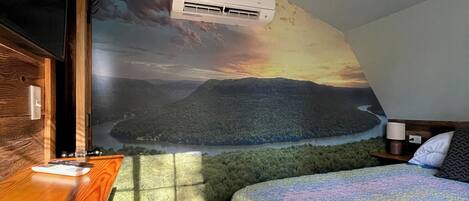 A bedroom with a mural of a landscape, a bed, a nightstand, a lamp, and an air conditioning unit.