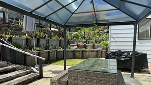 A covered patio with a glass table and benches, surrounded by a wooden fence and garden. - Enjoy our Wee Gem, she is over 120 years young and has been loving restored. (Albany)
