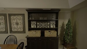 A dining area with a wooden table and chairs, a dark wooden buffet with decorative tiles and baskets, and a potted plant on a stand.