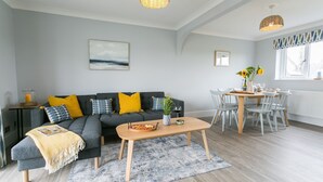 Een woonkamer met een grijze bank, gele kussens, een salontafel, en een eethoek met een tafel en stoelen.