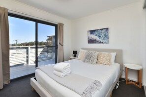 A modern bedroom with a large bed, a nightstand, a bedside table, and a view of a balcony with a railing.