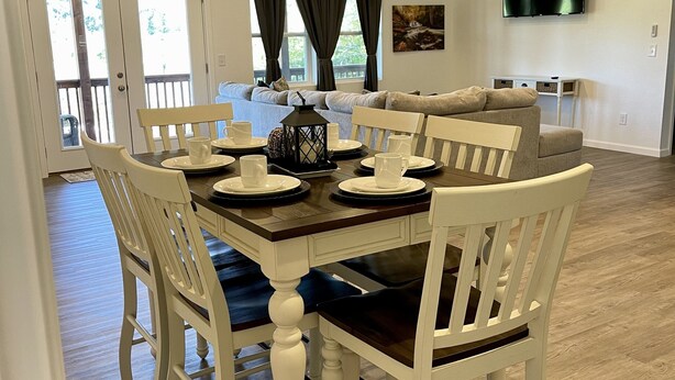 A dining area with a wooden table set for four, white chairs with dark cushions, a ceiling fan, and a TV mounted on the wall.