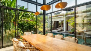 A spacious indoor area with a wooden table and wicker chairs, large windows, and a view of a swimming pool and greenery outside. - Villa Gaia Pererenan (Canggu)