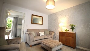 Un salon avec un canapé beige, un pouf à motifs écossais, un meuble en bois et un miroir au mur.