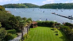 Vista costera con un puerto deportivo lleno de barcos, una zona verde con un pequeño edificio y colinas al fondo.