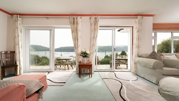Sala de estar con un amplio ventanal corredizo que da a un balcón con vistas a un lago y barcos. Hay un sofá, una mesita con un jarrón de flores y un banco.