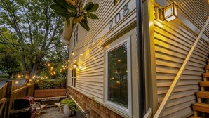 Une maison avec le numéro 208 sur le côté, une terrasse en bois avec des sièges d’extérieur et des guirlandes lumineuses.
