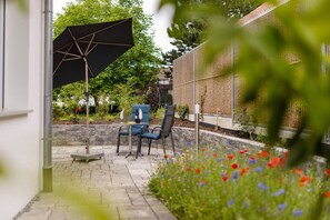 Un patio al aire libre con una mesa y sillas, un paraguas grande y un jardín con flores.