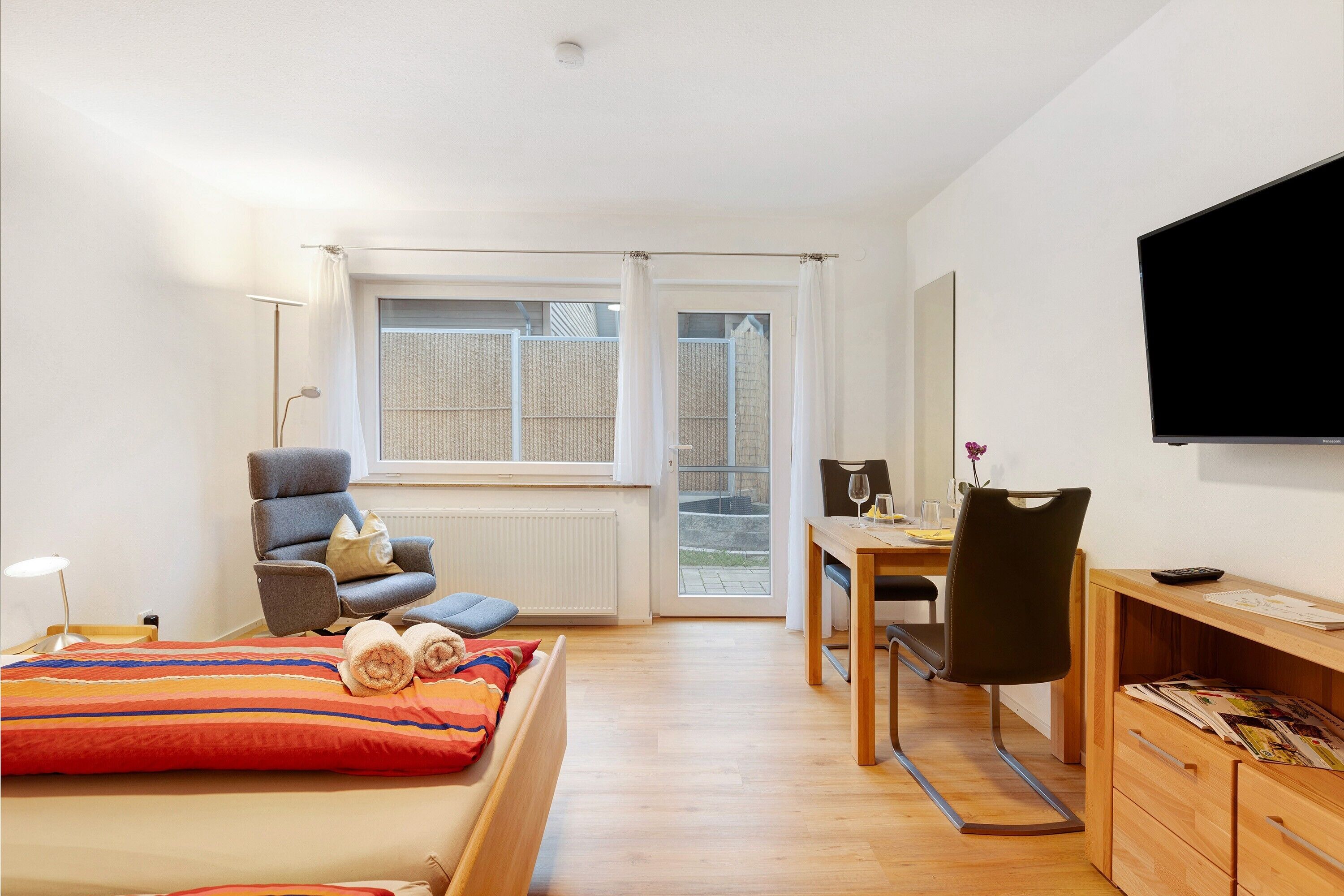 A bedroom with a bed, a desk, a chair, a TV, and a window.