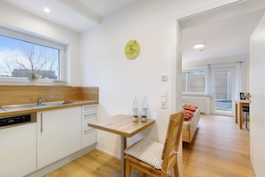 A modern kitchen with a dishwasher, a wooden table, and a chair. There is a clock on the wall and a window with a view outside.