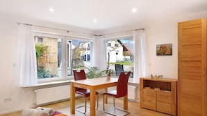 Une pièce comprenant une table à manger, des chaises, un lit avec une literie aux couleurs vives, une armoire en bois et de grandes fenêtres avec des rideaux.