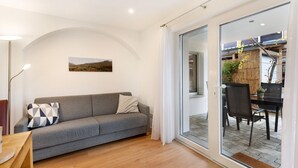 Un salon moderne avec un canapé gris, un lampadaire et une porte en verre donnant sur un patio extérieur aménagé avec une table et des chaises.