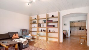 A living room with a sofa, coffee table, and a patterned rug. There are wooden shelves with various items and a kitchen visible through an archway. - Apartment 'Haus Steigstrasse' with Mountain View, Private Terrace and Wi-Fi (Opfenbach)