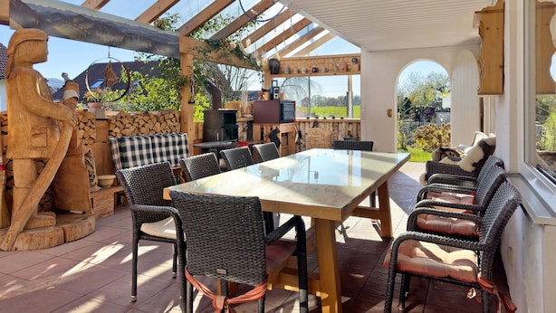 An outdoor patio with a glass-top table, wicker chairs, a wooden sculpture, and a stone wall. - Apartment 'Haus Steigstrasse' with Mountain View, Private Terrace and Wi-Fi (Opfenbach)