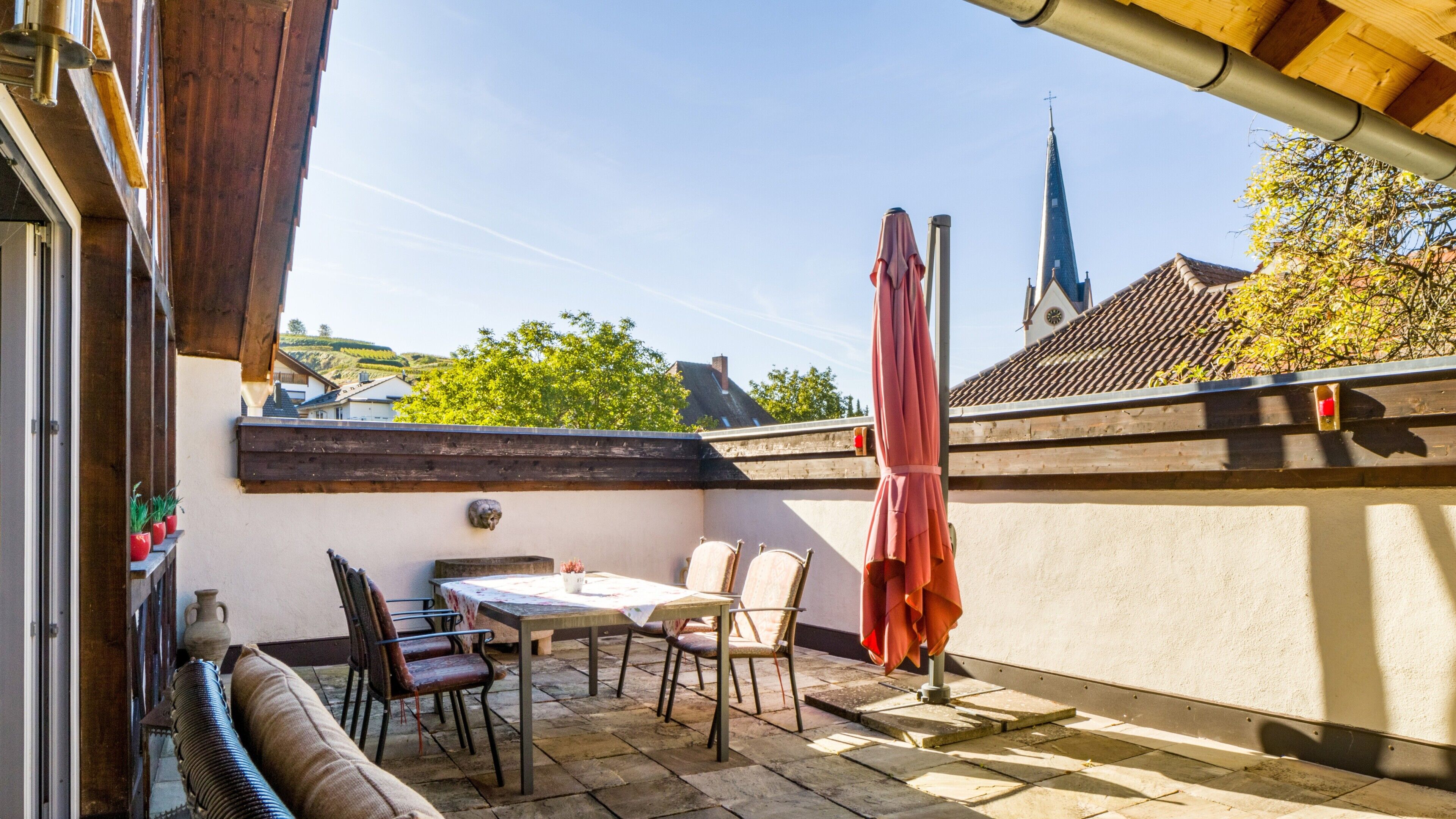A terrace with a table and chairs, a red umbrella, and a view of a church spire.
