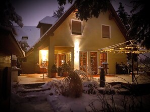 Ein Haus mit überdachter Terrasse, einem von Schnee bedeckten Garten und warmem Licht im Inneren.