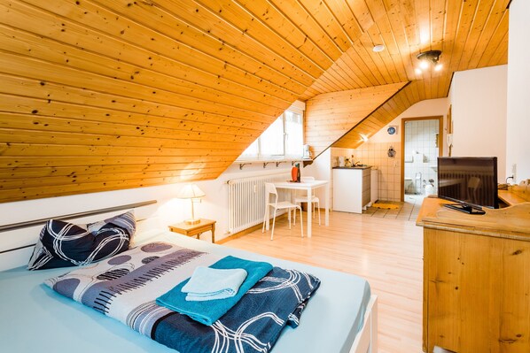 A cozy attic bedroom with a wooden ceiling, a bed with patterned bedding, a desk, and a kitchenette.