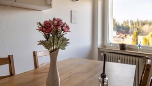 Uma mesa de jantar com um vaso de flores, um copo de água e uma janela com vista para árvores.